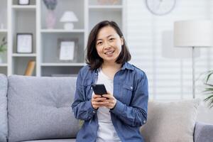 Porträt von ein jung schön asiatisch Frau beim Zuhause auf das Couch, Frau lächelnd und suchen beim das Kamera halten ein Telefon, Smartphone Anwendung Benutzer, zufrieden und glücklich. foto