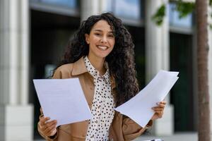 elegant jung spanisch Geschäftsfrau halten Papiere und lächelnd, Stehen draußen ein Büro Gebäude auf ein sonnig Tag. foto