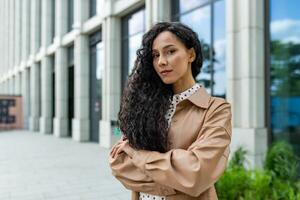 ein jung Fachmann Frau steht selbstbewusst draußen ein modern Büro Gebäude, ausströmend Stärke und Ambition im ein korporativ Einstellung. foto