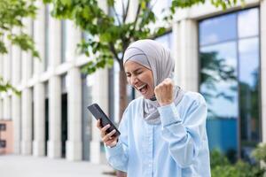 froh Frau im Hijab Gehen im das Stadt, empfangen Benachrichtigung von online Sieg und gut Nachrichten auf Telefon, Geschäft Frau draußen Büro Gebäude, halten Hand oben Triumph Gewinner Geste. foto