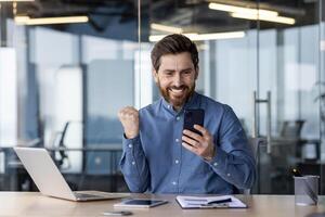 triumphierend männlich Unternehmer freut sich im ein Geschäft Erfolg beim seine modern Arbeitsplatz mit Smartphone und Laptop. foto