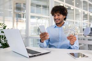 froh männlich Unternehmer im ein modern Büro Umfeld, halten ein Handy, Mobiltelefon Telefon und mehrere uns Dollar Rechnungen, lächelnd und suchen beim Telefon. foto