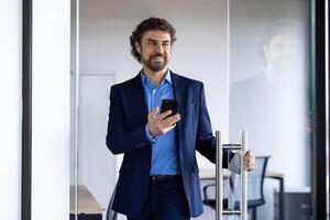 erfolgreich froh Geschäftsmann öffnet Glas Büro Tür, Mann mit Telefon im Hände lächelnd, zufrieden mit Leistung Ergebnisse, Boss im Geschäft Anzug. foto