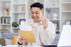 glücklich und erfolgreich asiatisch empfangen Mail Benachrichtigung Briefumschlag mit gut Nachricht, Mann Sitzung beim Tabelle beim heim, Arbeiten im Zuhause Büro im Leben Zimmer, freudig feiern gewinnen Nachricht. foto