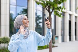 schön arabisch Geschäftsfrau im Hijab mit Handy, Mobiltelefon Telefon zum Forderung, glücklich winken beim das Bildschirm draußen. foto