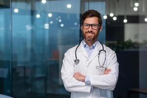 Porträt von reifen Arzt mit Bart, Mann im Weiß medizinisch Mantel lächelnd und suchen beim Kamera mit gekreuzt Waffen Arbeiten Innerhalb modern Klinik. foto