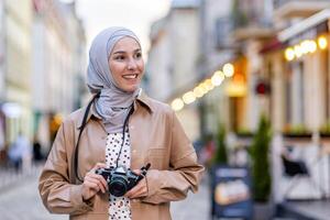 jung schön Frau Gehen im das Abend Stadt im Kopftuch, Tourist mit Kamera und tragen ein Hut inspiziert das historisch Stadt lächelnd mit Zufriedenheit, Muslim Frau auf ein Reise. foto