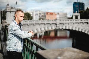ein Mann im ein Denim Jacke steht auf ein Brücke mit Blick auf ein Fluss foto