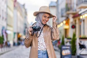 Porträt von jung schön Muslim Frau im Hijab und Hut, Tourist Gehen im Abend Stadt mit Kamera, Frau Reisen zu anders Länder. foto