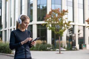 ein konzentriert Frau genießen ihr Wiedergabeliste während Handhabung ihr Smartphone im ein gemächlich Haltung, gekleidet im Fitness Kleidung, mit modern die Architektur und Grün im das Hintergrund. foto