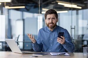 verwirrt jung Fachmann mit ein Bart im ein Blau Hemd suchen beim seine Smartphone, ausdrücken Verwechslung im ein zeitgenössisch Büro Einstellung. foto