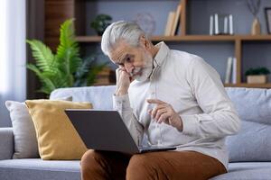 konzentriert Alten Mann mit Weiß Haar und Bart tief vertieft im mit ein Laptop während Sitzung auf ein Sofa im ein gut beleuchtet Leben Zimmer, abbilden modern Technologie verwenden durch Senioren. foto