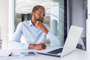 Denken ernst afrikanisch amerikanisch Geschäftsmann Arbeiten Innerhalb Büro, erfolgreich Geschäft Mann Boss suchen beim Laptop Bildschirm Nachdenken wichtig technisch Entscheidung. foto