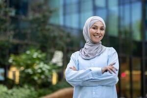 Porträt von ein heiter Muslim Frau mit Kopftuch, selbstbewusst Stehen mit gekreuzt Waffen im ein modern Stadt Umfeld. foto