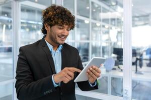 jung erfolgreich Geschäftsmann mit Tablette Computer , lächelnd spanisch Mann Arbeiten Innerhalb ein modern Büro Gebäude beim Arbeit foto