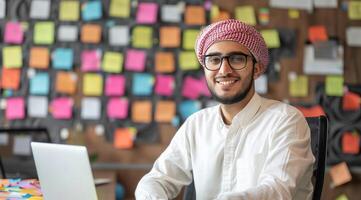 Porträt von ein lächelnd jung arabisch Mann Sitzung beim ein Schreibtisch mit ein Laptop im ein modern Büro foto