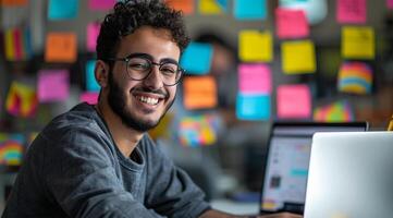 Porträt von ein lächelnd jung arabisch Mann Sitzung beim ein Schreibtisch mit ein Laptop im ein modern Büro foto