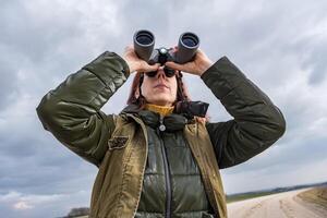 weiblich Ornithologe Vogelmann oder Forscher Uhren Vögel mit Fernglas gegen ein Hintergrund von ein stürmisch Himmel foto