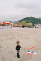 wenig Mädchen steht auf das Strand mit ein Spule von ein mehrfarbig Drachen auf das Sand und sieht aus beim Es. zurück Aussicht foto