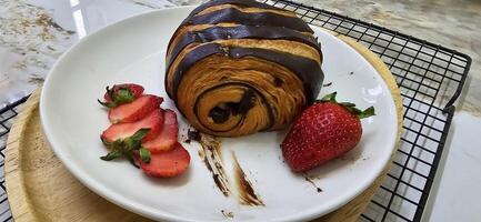 frisch hausgemacht gestreift Schokolade Croissant mit Schokolade Füllung auf ein runden Weiß Platte, serviert mit frisch Erdbeere foto