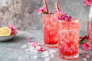 zwei erfrischend Rosa Cocktails geschmückt mit Blumen im Kristall Gläser, perfekt zum Sommer- Parteien oder Valentinsgrüße Tag Feierlichkeiten, mit verstreut Eis auf ein texturiert Hintergrund foto