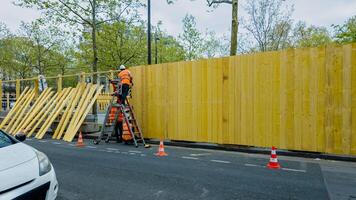 Konstruktion Arbeiter im hoch Sichtweite Weste montiert hölzern Zaun durch das Straßenrand, Signalisierung städtisch Entwicklung und Infrastruktur Upgrades, Sicherheit beim Arbeit Konzept foto