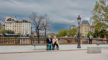 Alten Paar Sitzung auf ein Pariser Bank mit ein Koffer, umgeben durch städtisch Landschaft, bedeuten Reise und Pensionierung Freizeit, Paris, Frankreich, April 14, 2024 foto