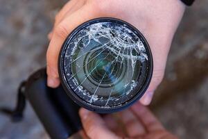 Hände von jung Fotograf halten dslr Foto Kamera mit gebrochen Linse Filter Glas nach wenn fallen Nieder auf zu das Boden. schließen hoch. zerstört geknackt Foto Filter. Makro. oben Sicht. selektiv Fokus.