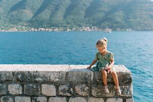 wenig Mädchen mit ein Sanft Spielzeug sitzt auf ein Stein Zaun auf das Strand und Picks es mit ihr Finger foto