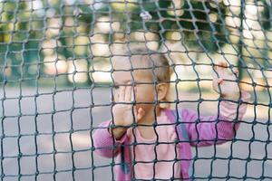 wenig Mädchen steht hinter ein Tennis Netz und ruht ihr Hände auf es foto