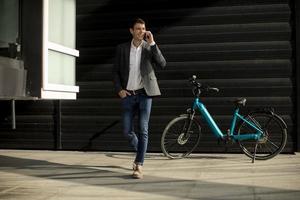 junger Geschäftsmann, der vom E-Bike weggeht und Handy benutzt foto