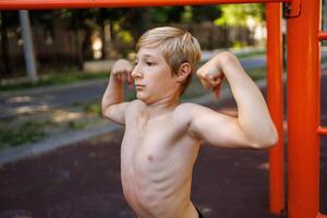 ein Teenager mit ein sportlich bauen zeigt an aus seine Muskulatur. Straße trainieren auf ein horizontal Bar im das Schule Park. foto