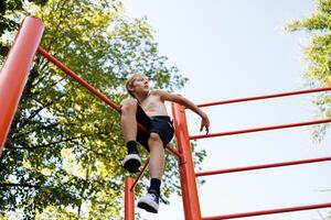 ein Teenager Junge kletterte oben das Gymnastik- Querlatte sitzt und ruht. Straße trainieren auf ein horizontal Bar im das Schule Park. foto