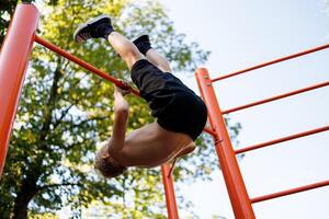 Unterseite Aussicht von ein Teenager durchführen Gymnastik- Übungen. Straße trainieren auf ein horizontal Bar im das Schule Park. foto