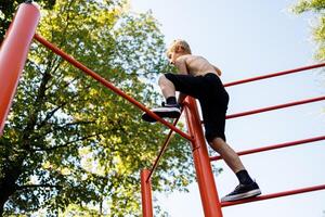 Unterseite Aussicht von ein Teenager Klettern auf ein Gymnastik Bar. Straße trainieren auf ein horizontal Bar im das Schule Park. foto