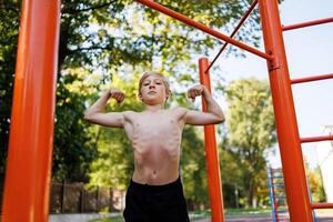 ein Teenager Junge von ein sportlich bauen zeigt an seine Muskeln. Straße trainieren auf ein horizontal Bar im das Schule Park. foto