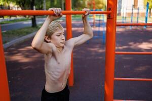 ein Teenager mit sportlich Muskel Entwicklung hält auf zu ein Bar. Straße trainieren auf ein horizontal Bar im das Schule Park. foto