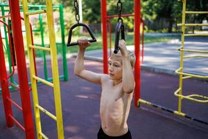 ein Teenager mit ein sportlich bauen hält auf zu das Handläufe von ein Sport Ausrüstung. Straße trainieren auf ein horizontal Bar im das Schule Park. foto