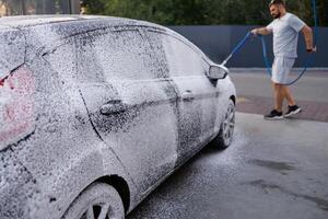 das Seite Türen von das Auto sind bedeckt mit Schaum, das Mann im das Hintergrund ist bewirbt sich Schaum zu das Wagen. ein Auto beim ein selbst Bedienung Auto waschen. foto