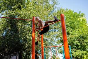 ein Junge Athlet klettert das Querlatte zu ausführen ein formal Element. Straße trainieren auf ein horizontal Bar im das Schule Park. foto