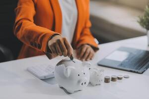 schön asiatisch Geschäftsfrau einfügen Münzen im ein Stift Schweinchen Bank auf das Tisch. Speichern Geld, Investition, Währung Konzept foto