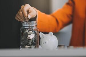 schön asiatisch Geschäftsfrau einfügen Münzen im ein Stift Schweinchen Bank auf das Tisch. Speichern Geld, Investition, Währung Konzept foto