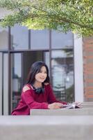 Porträt von schön sexy asiatisch Frau erkunden Cafe draußen. Mädchen Sitzung entspannend lesen ein Buch suchen beim das Kamera zum Inhalt oder Werbeaktionen foto