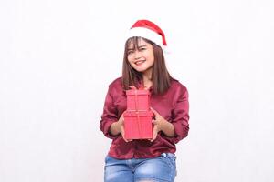 schön jung asiatisch indonesisch Mädchen lächelnd bringen Geschenk Box beim Weihnachten Santa claus Hut modern rot Hemd Outfit geben Geschenke heiter Geschenke auf Weiß Hintergrund zum Beförderung und Werbung foto