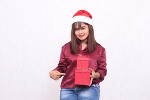 schön jung asiatisch indonesisch Mädchen freudig Tragen Geschenk Box im Weihnachten Santa claus Hut modern rot Hemd Outfit spitz Seite Box auf Weiß Hintergrund zum Beförderung und Werbung foto