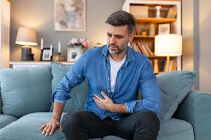 schreckliche bauchschmerzen. frustrierter gutaussehender junger mann, der seinen bauch umarmt und die augen geschlossen hält, während er zu hause auf der couch liegt foto