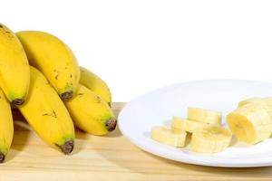 Bündel Bananen auf Holztisch neben Teller mit geschnittenen Früchten, weißer Hintergrund