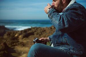bärtig Mann Trinken heiß Tee oder Kaffee während nehmen ein Kaffee brechen, ruhen durch Ozean. brechen Wellen auf das Hintergrund foto