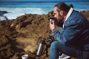 männlich Reisender gießt heiß Tee von Thermosflasche Flasche in Becher im das felsig Strand. Personen. Natur. Tourismus und Camping Konzept foto