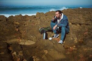 entspannt Tourist mit Thermosflasche, haben ausruhen, genießt seine Kaffee brechen, Sitzung mit seine Augen geschlossen auf das felsig Cliff durch Meer foto